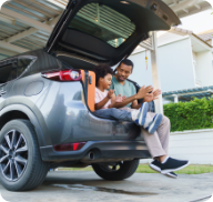 A father and his young son sit in the open hatch of an SUV parked in a garage.  